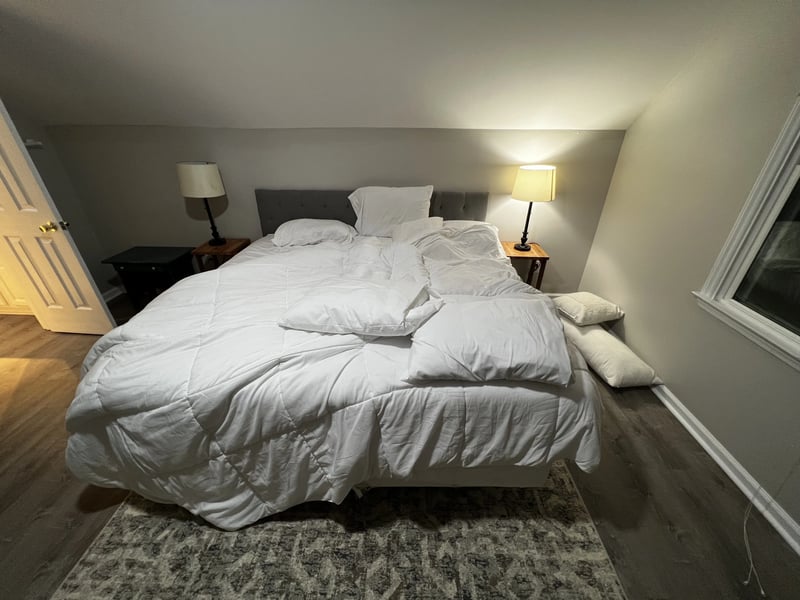 An unmade bed with rumpled white bedding and scattered pillows in a bedroom with a gray headboard