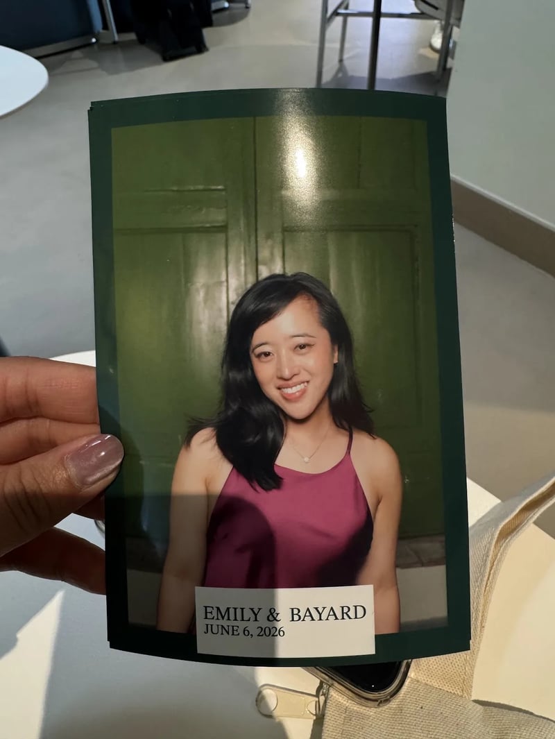 A hand holds a printed photobooth portrait of a smiling woman in a magenta top in front of green doors